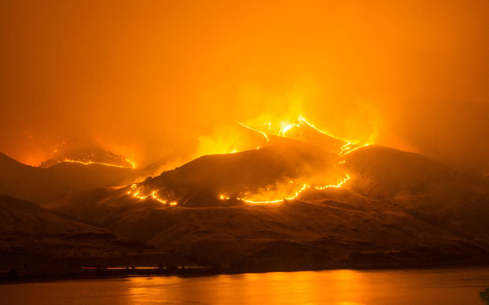 A wildfire burning along dry hills at night, reflected in a calm river