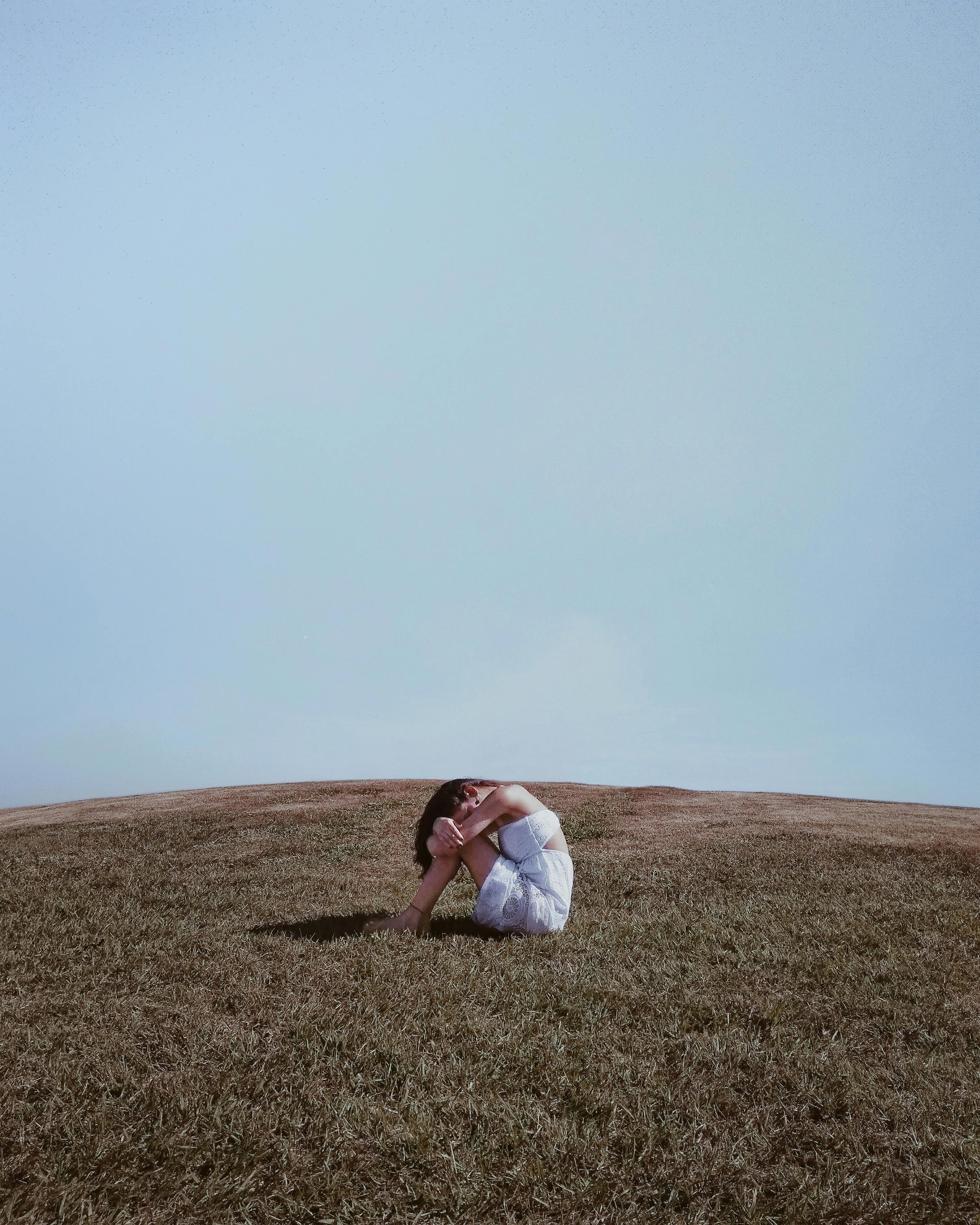 一个女人穿着白色连衣裙，独自坐在明亮的天空下的草山上