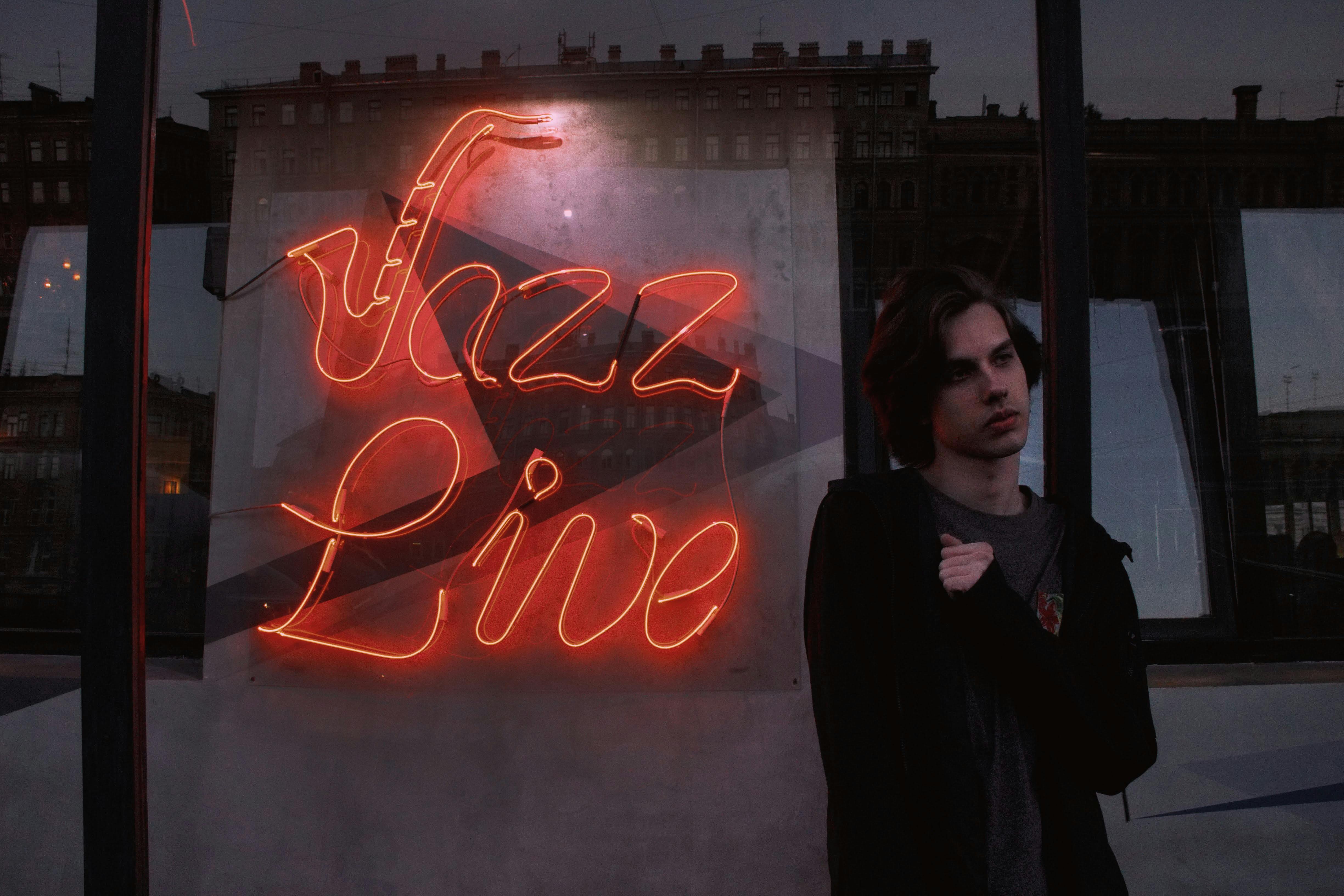 A young adult standing by a glowing neon 'Jazz Live' sign, reflecting the urban vibe at night.