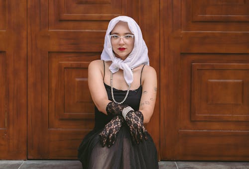 Young Woman Wearing a Black Dress, Lace Gloves and White Headscarf Sitting in front of Wooden Door 