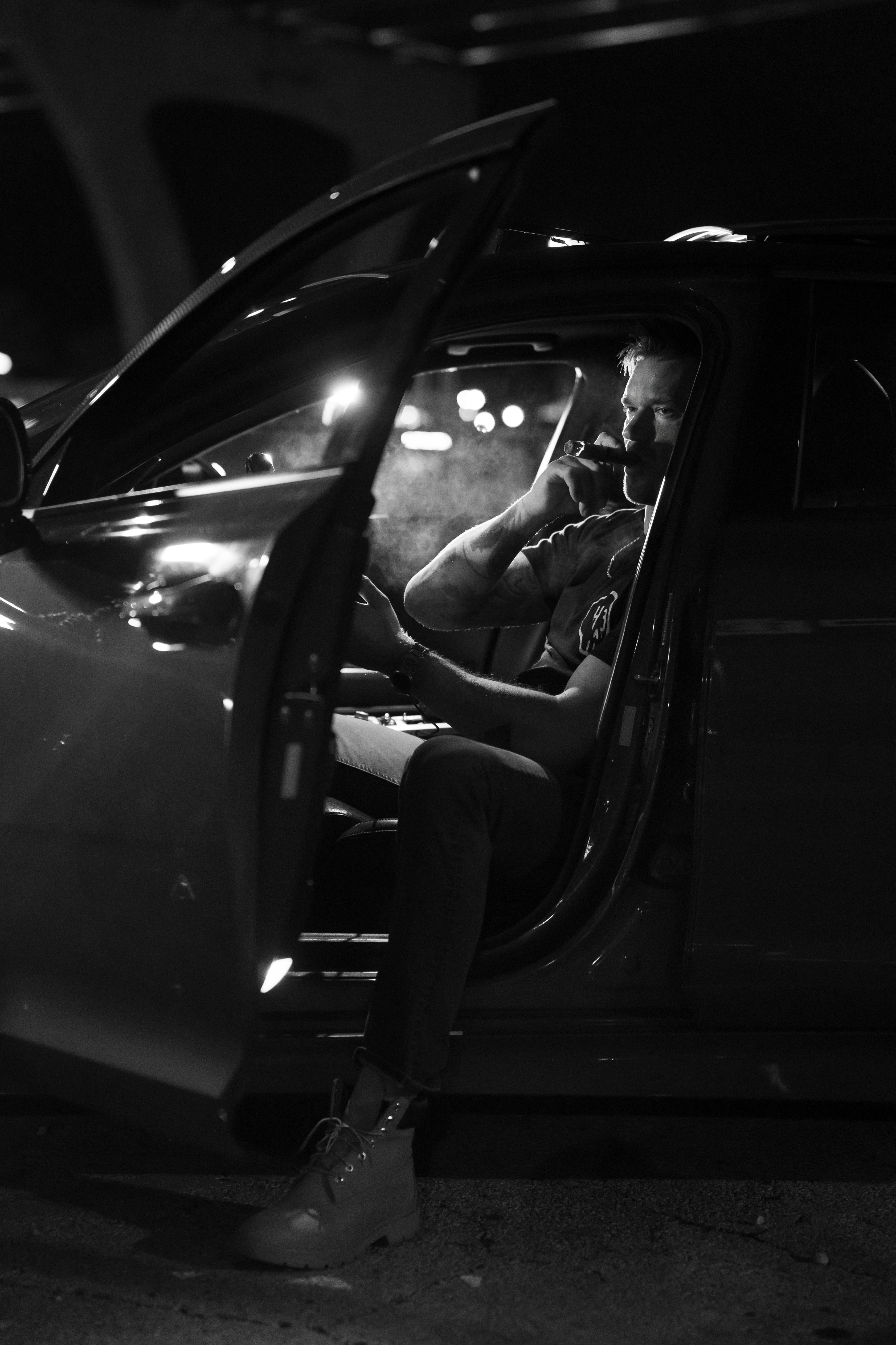 a man sitting in his car with doors open smoking a cigarette at night black and white