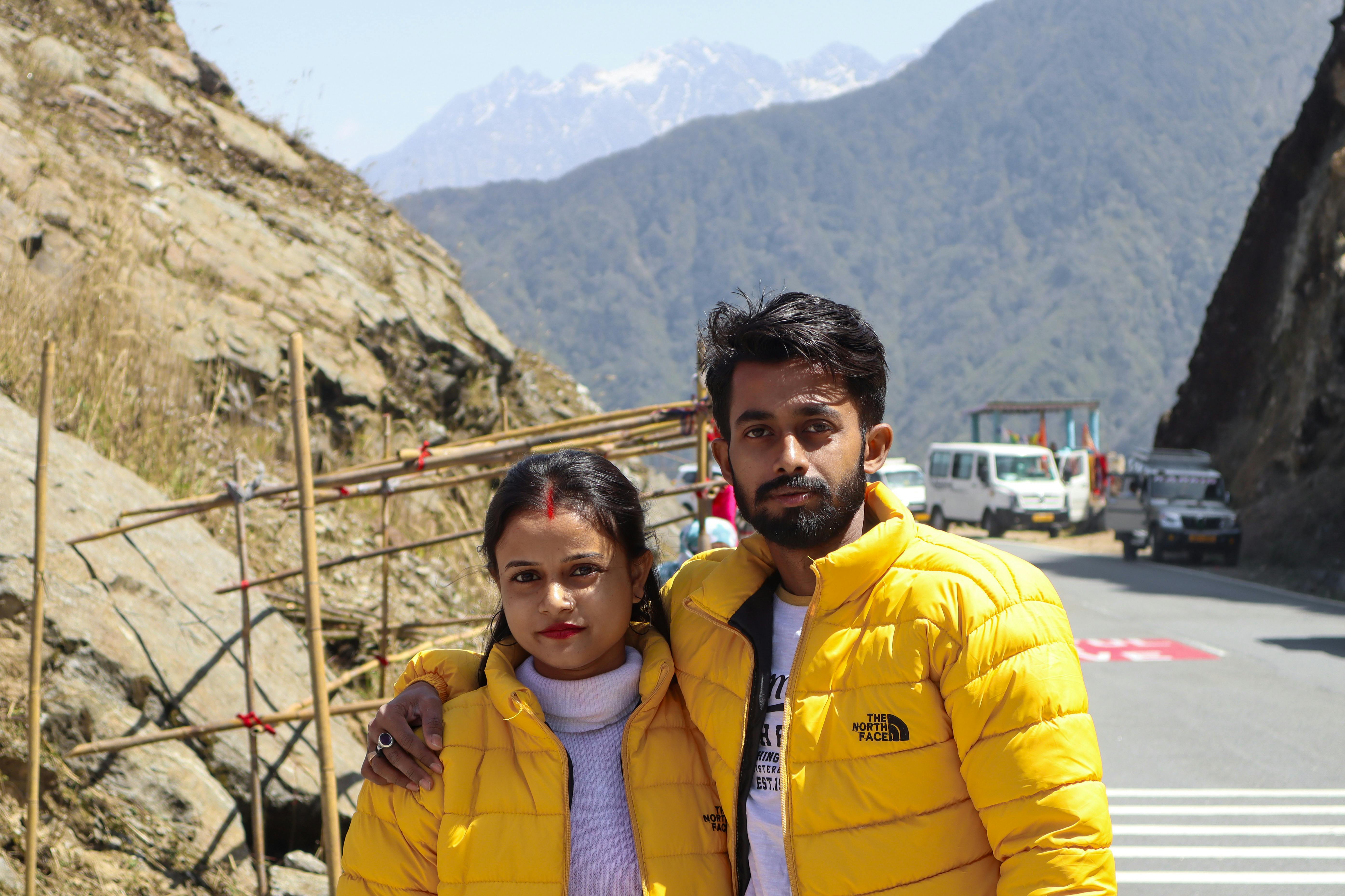 Couple wearing yellow jackets posing on a mountainous road with cars and rocky terrain.