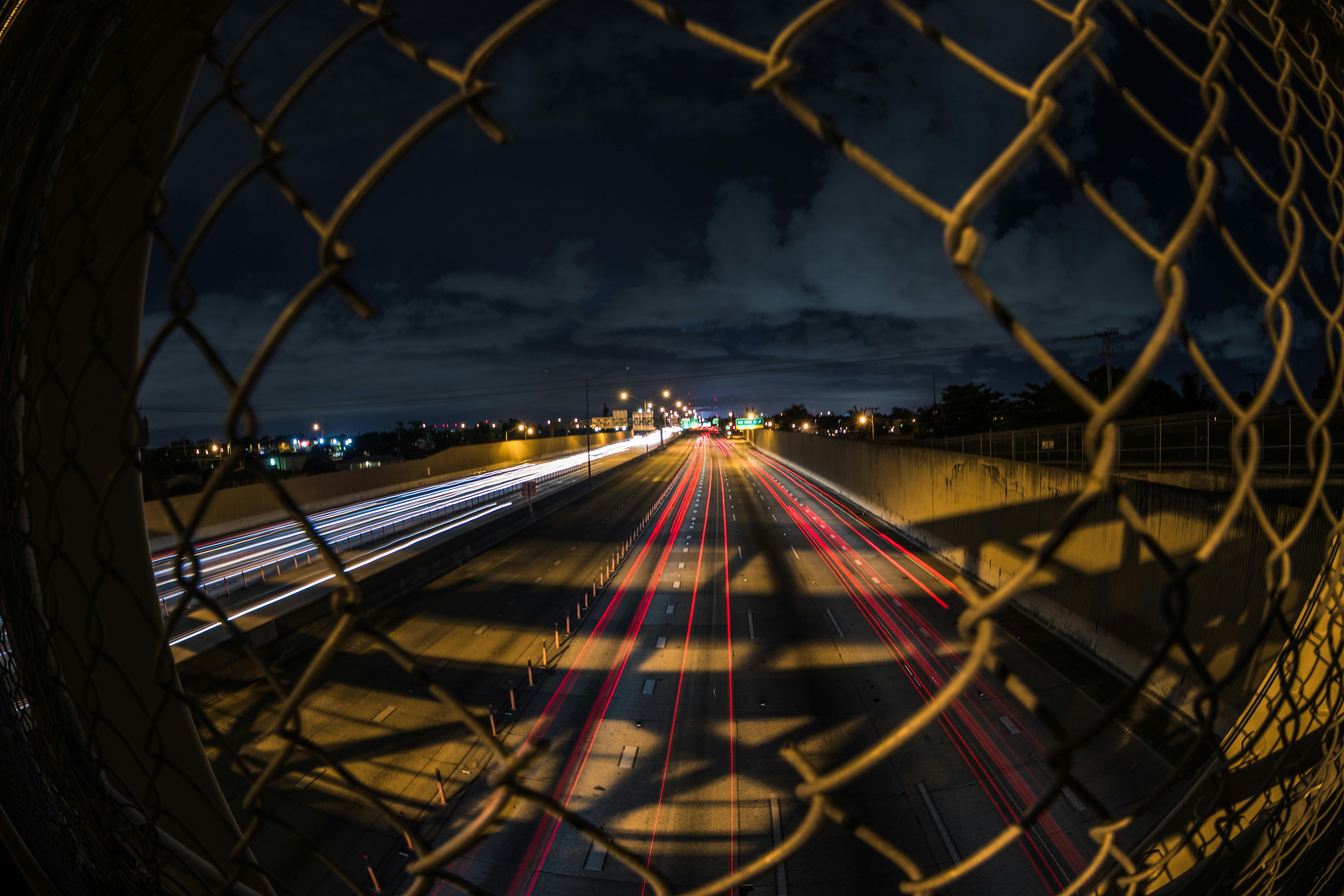 long exposure of cars on a street at night photographed through a hole in a fence