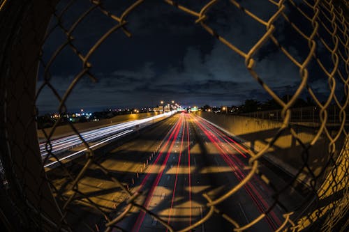 Long Exposure of Cars on a Street at Night Photographed through a Hole in a Fence 