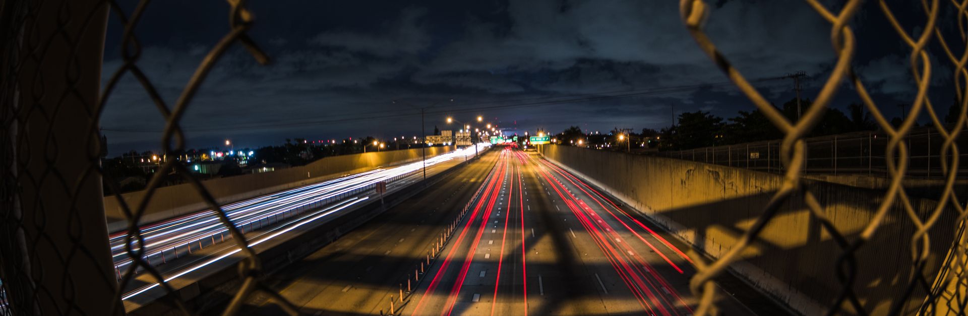 A long exposure shot capturing light trails on a Miami highway at night through a fence.