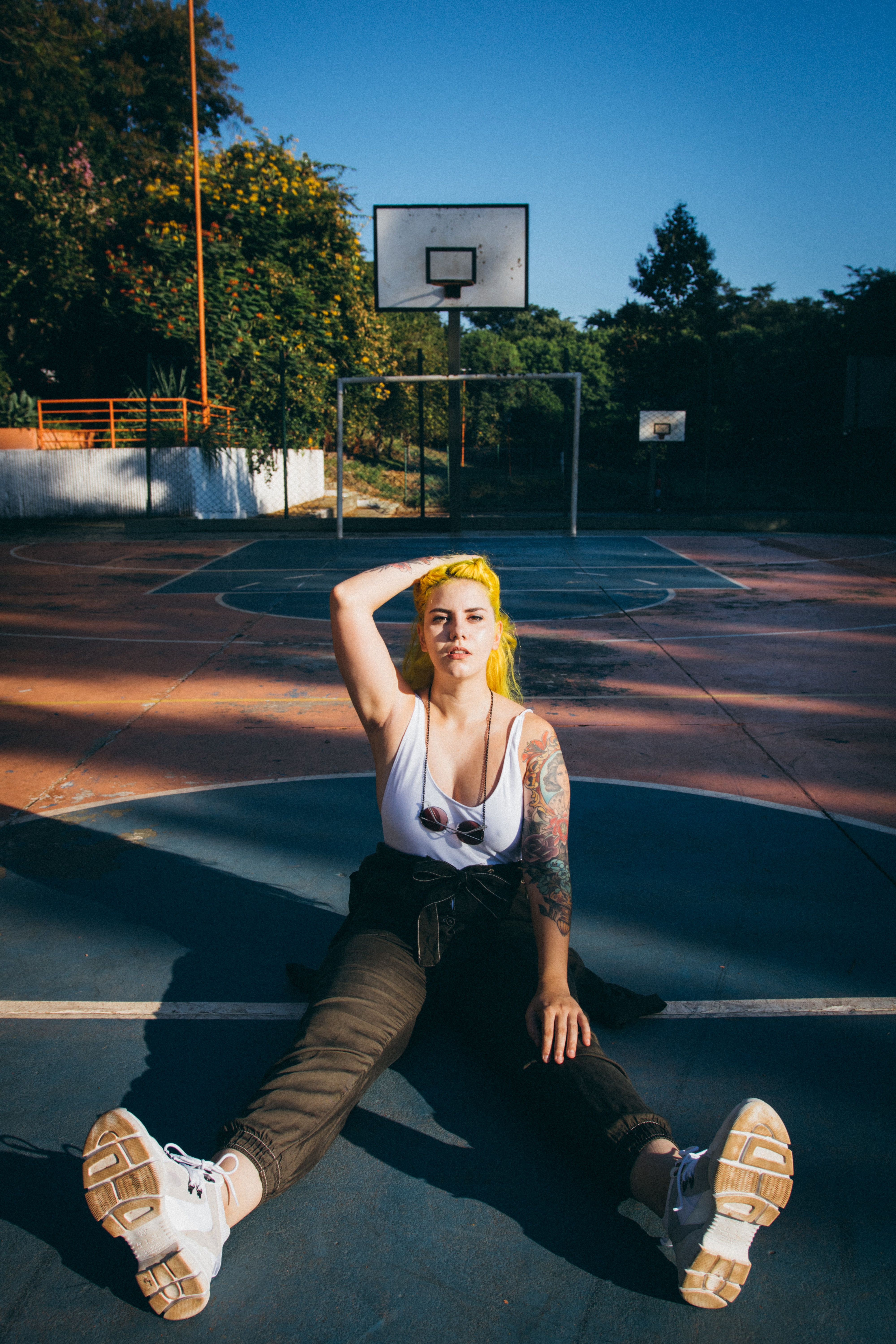 A woman with yellow hair sitting on an outdoor basketball court in the sun, showcasing a casual style.