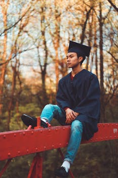 一位年轻的毕业生头戴帽子、身着长袍，坐在户外的金属梁上，四周树木环绕