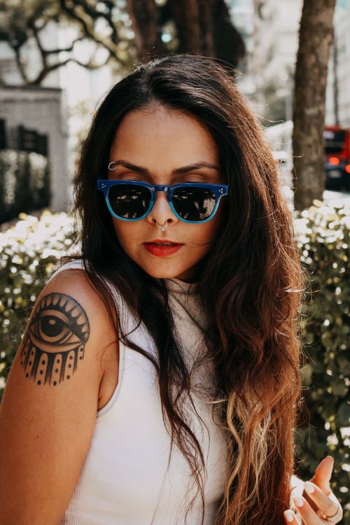Free Trendy woman poses outdoors with sunglasses, a white top, and an eye-catching arm tattoo. Stock Photo