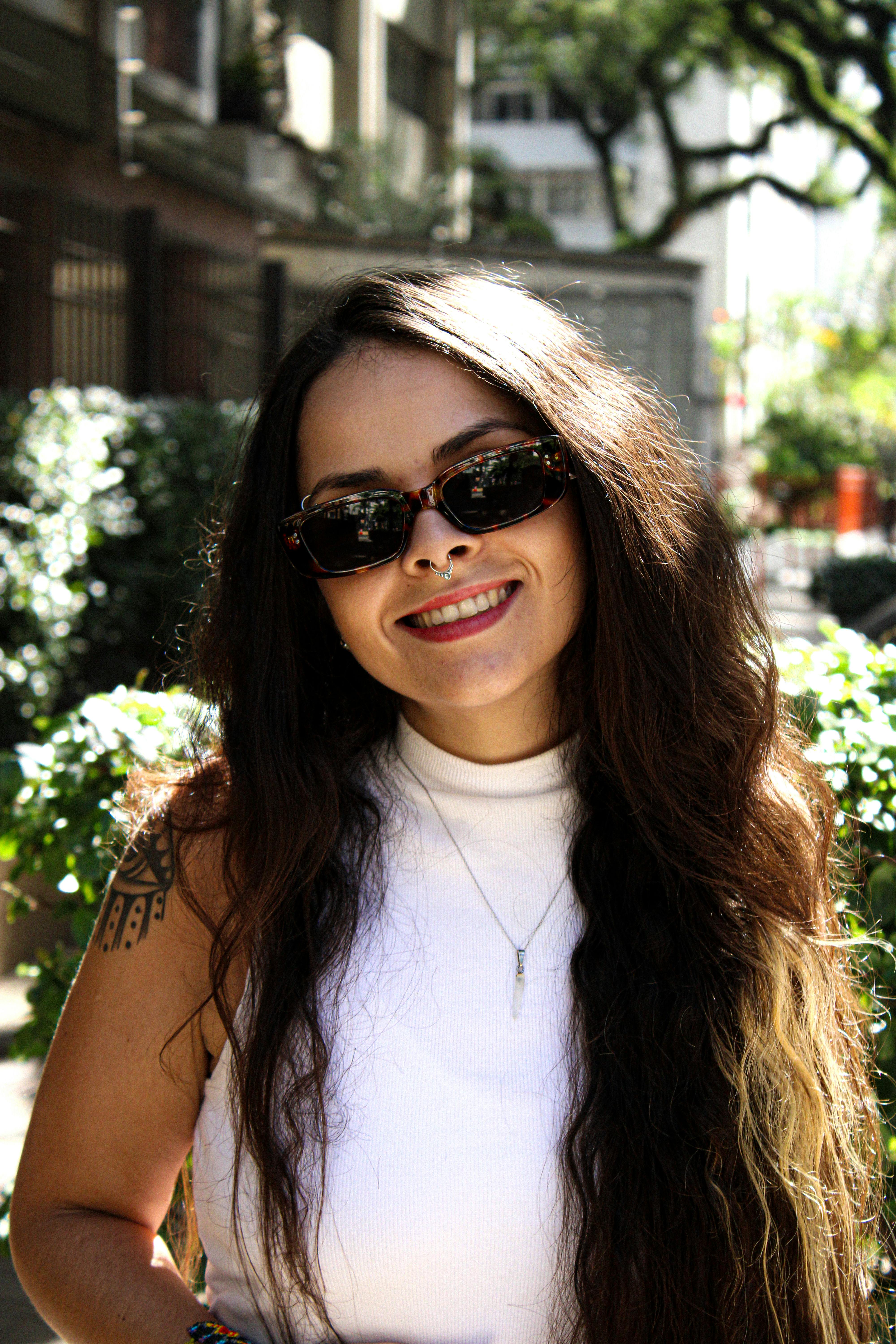A young woman with sunglasses and long hair smiles brightly outdoors in a sunny setting.