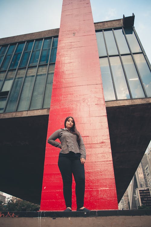 Woman in Gray Long Sleeve Shirt and Black Pants Standing Beside Red Brick Wall