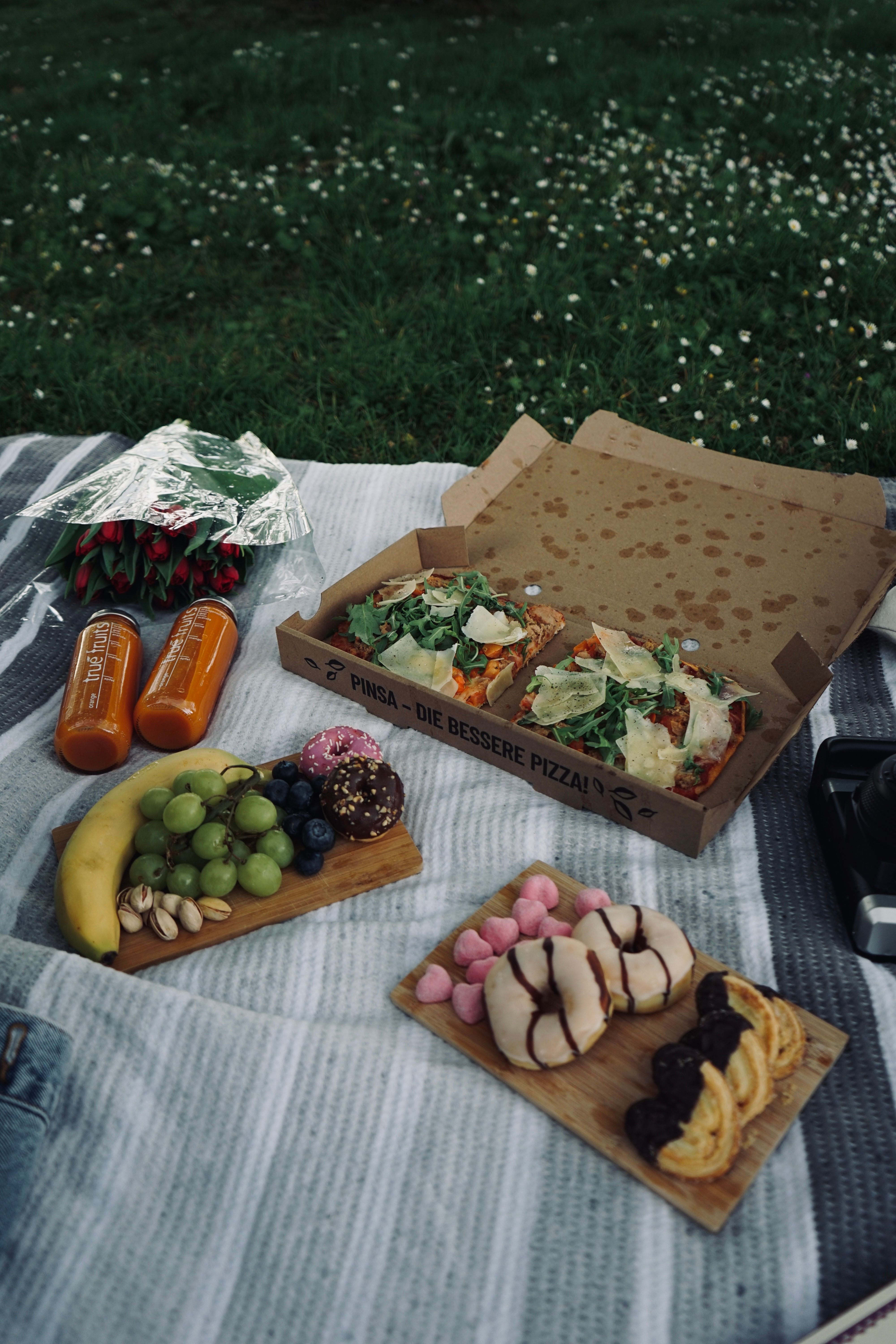 A delicious picnic spread featuring pinsa, fruits, donuts, and juice bottles on a blanket outdoors.