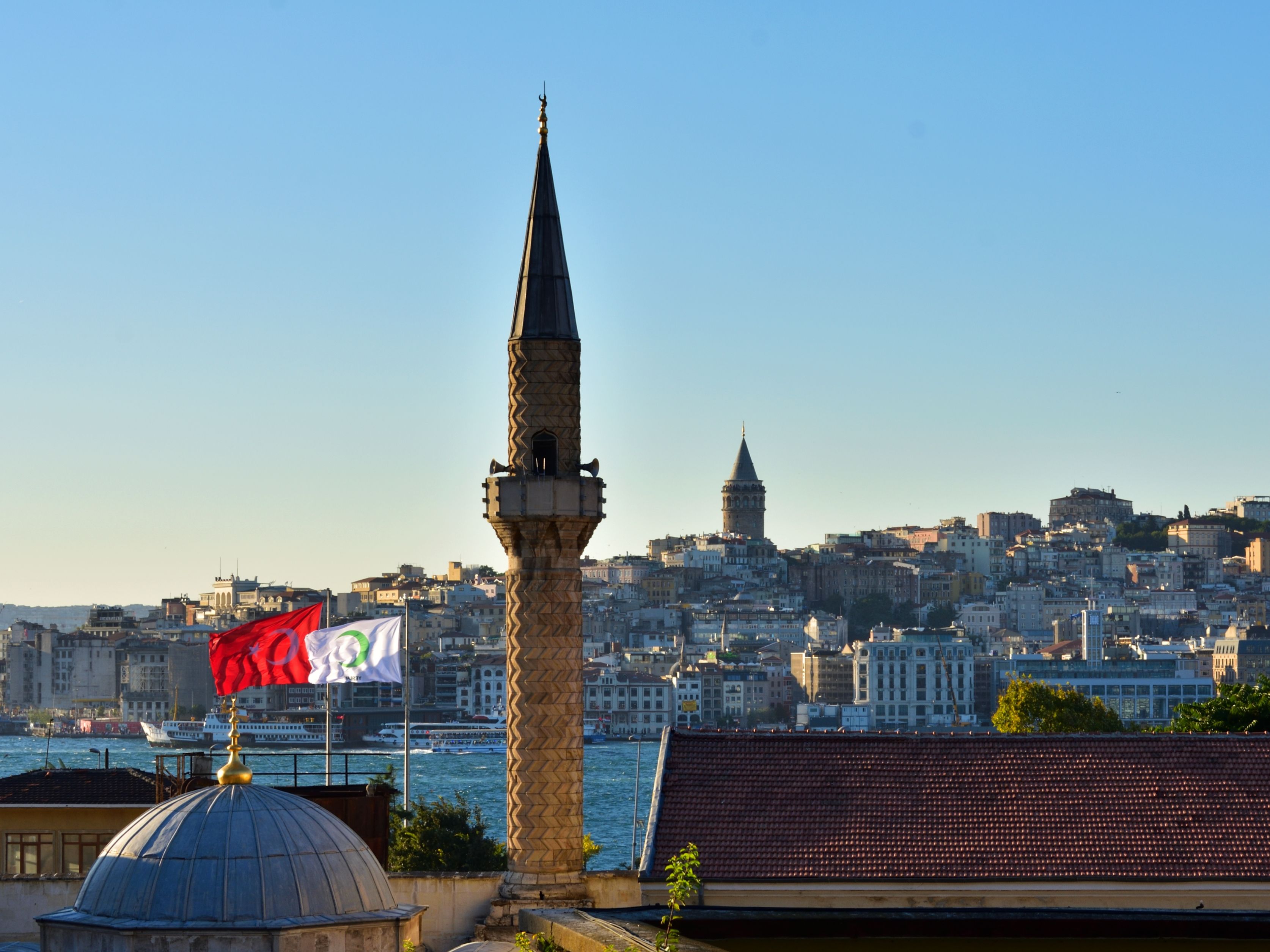 Vue d'Istanbul montrant un minaret au premier plan, la tour de Galata au loin et un drapeau turc flottant près de l'eau.