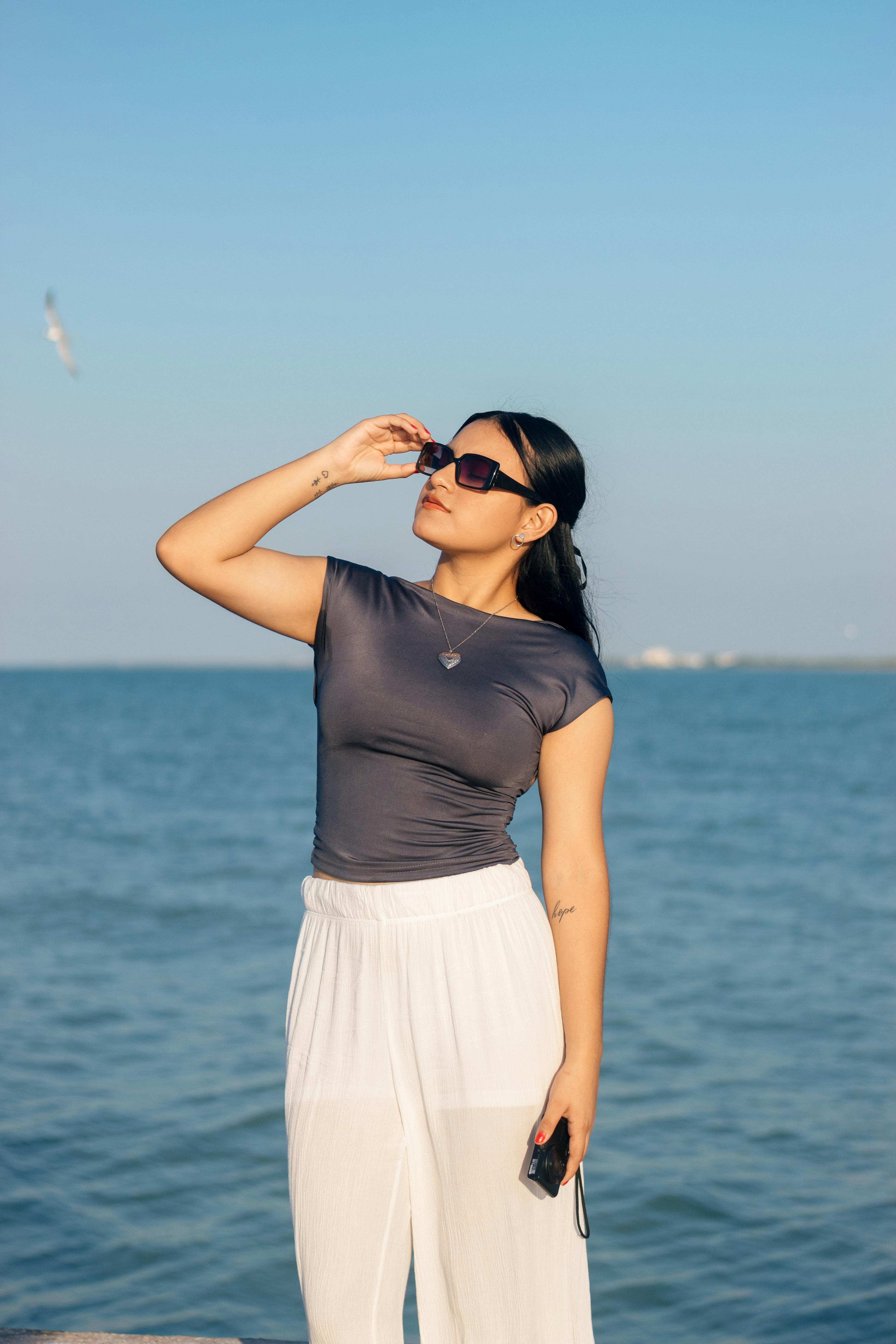 Stylish young woman wearing sunglasses poses by the sea under a clear, blue sky.
