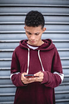 A young man in a hoodie texting on his smartphone outdoors against a roller shutter backdrop.