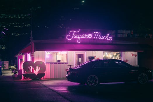 A neon sign illuminates a taqueria with a parked car at night, capturing urban nightlife.