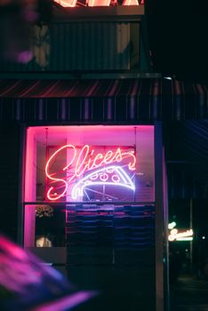 Neon pizza sign glowing brightly at night, outside a pizzeria offering slices.