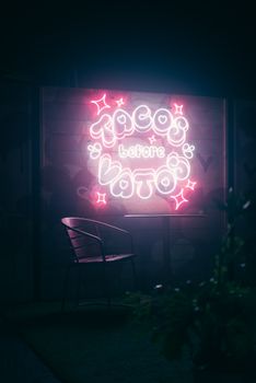 Vibrant neon sign reading 'Tacos Before Vatos' glowing in the night with a vintage chair beside it.