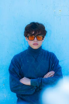 Young man with sunglasses posing against a blue wall, showcasing modern fashion style.