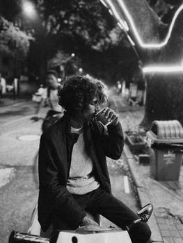 A man sitting on a motorbike, sipping coffee on a city street at night.