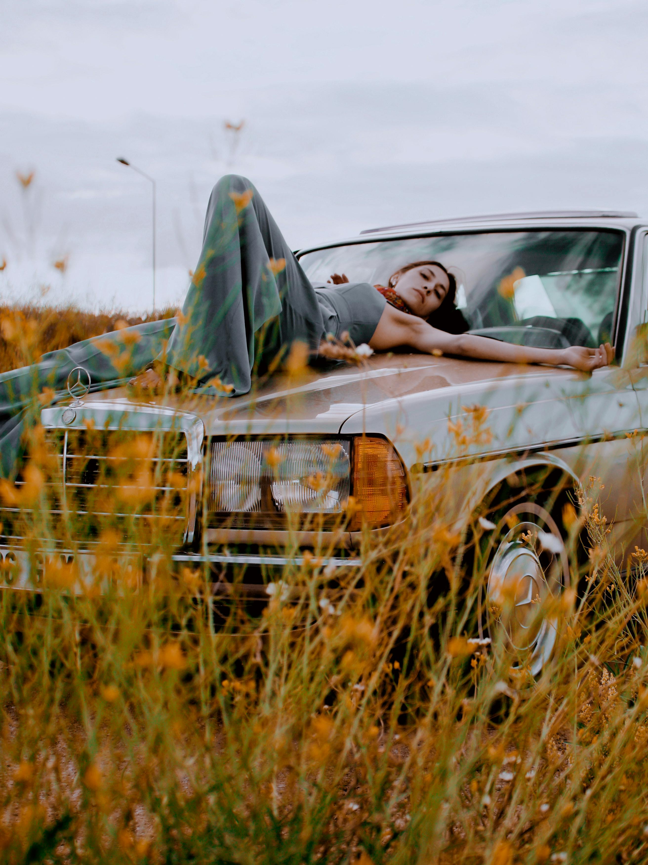 Young woman in baggy pants posing on a vintage car surrounded by wildflowers in a serene outdoor setting.