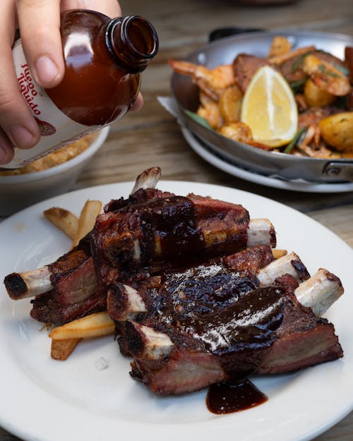 A person pouring sauce on ribs and fries