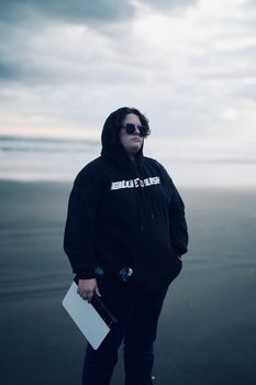 Stylish person in sunglasses and black hoodie on a moody beach. Perfect for fashion and lifestyle imagery.