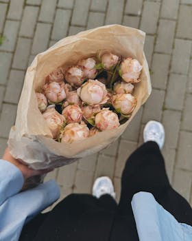 A person carries a bouquet of pink roses wrapped in kraft paper while walking on a city sidewalk.