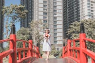 一位身着白色连衣裙的女子，手持花束，站在红色的桥上，背景是摩天大楼