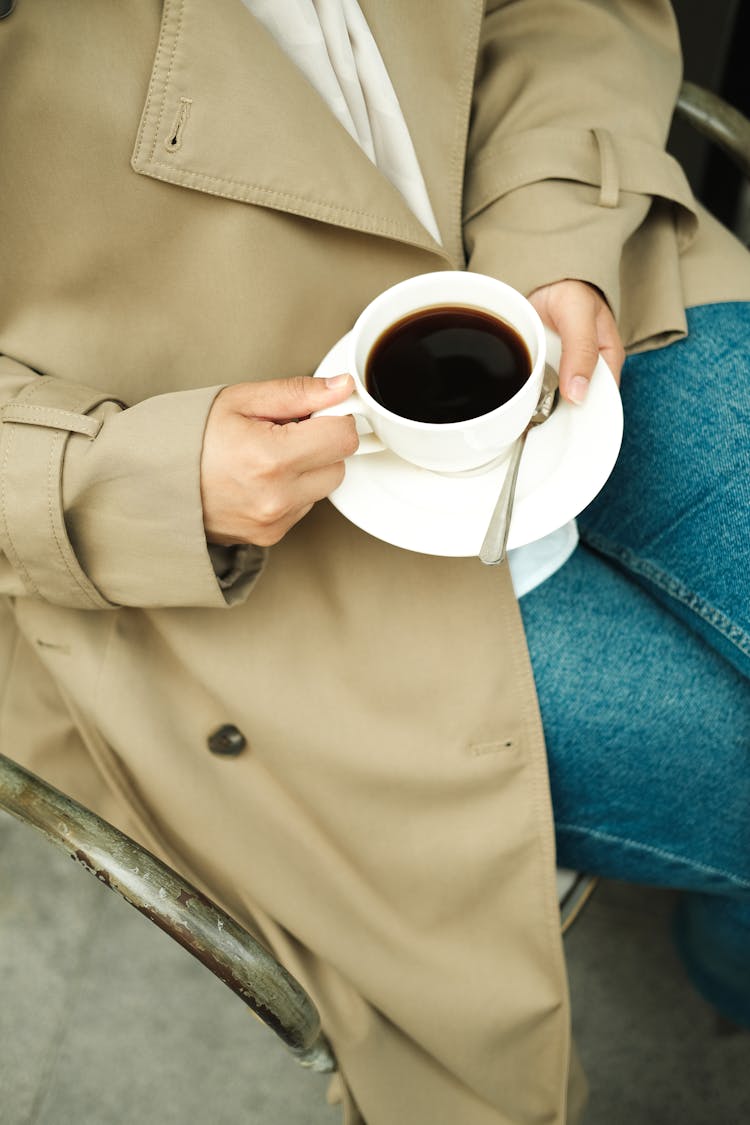 Close-up Of Woman In Jeans And A Trench Sitting And Holding A Cup Of Coffee