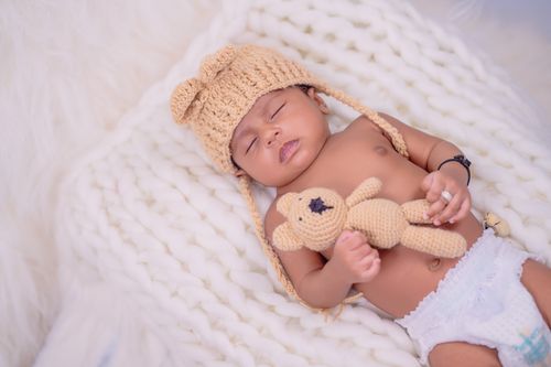 Free A peaceful baby sleeping on a cozy white blanket, wearing a knitted hat and holding a crocheted bear toy. Stock Photo
