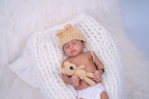 Free Charming indoor studio shot of a sleeping baby wearing a knitted hat and holding a stuffed teddy bear. Stock Photo