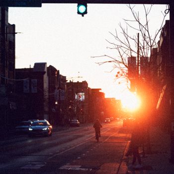 A cyclist rides through a city street during sunset, with cars and urban landscape in view.