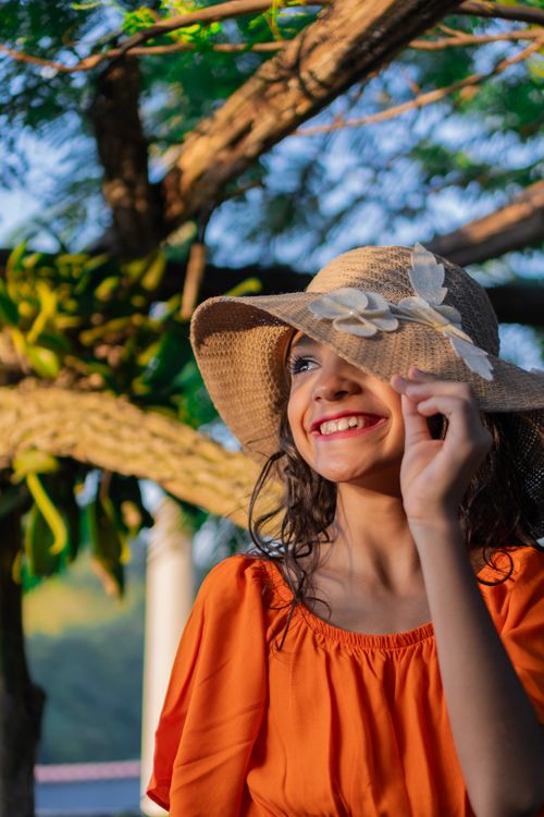 Free A cheerful girl in an orange dress and sunhat smiling under a tree on a sunny day. Stock Photo