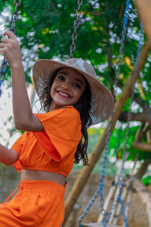 Free Joyful young girl with wide-brimmed hat enjoying a swing against a lush green backdrop. Stock Photo
