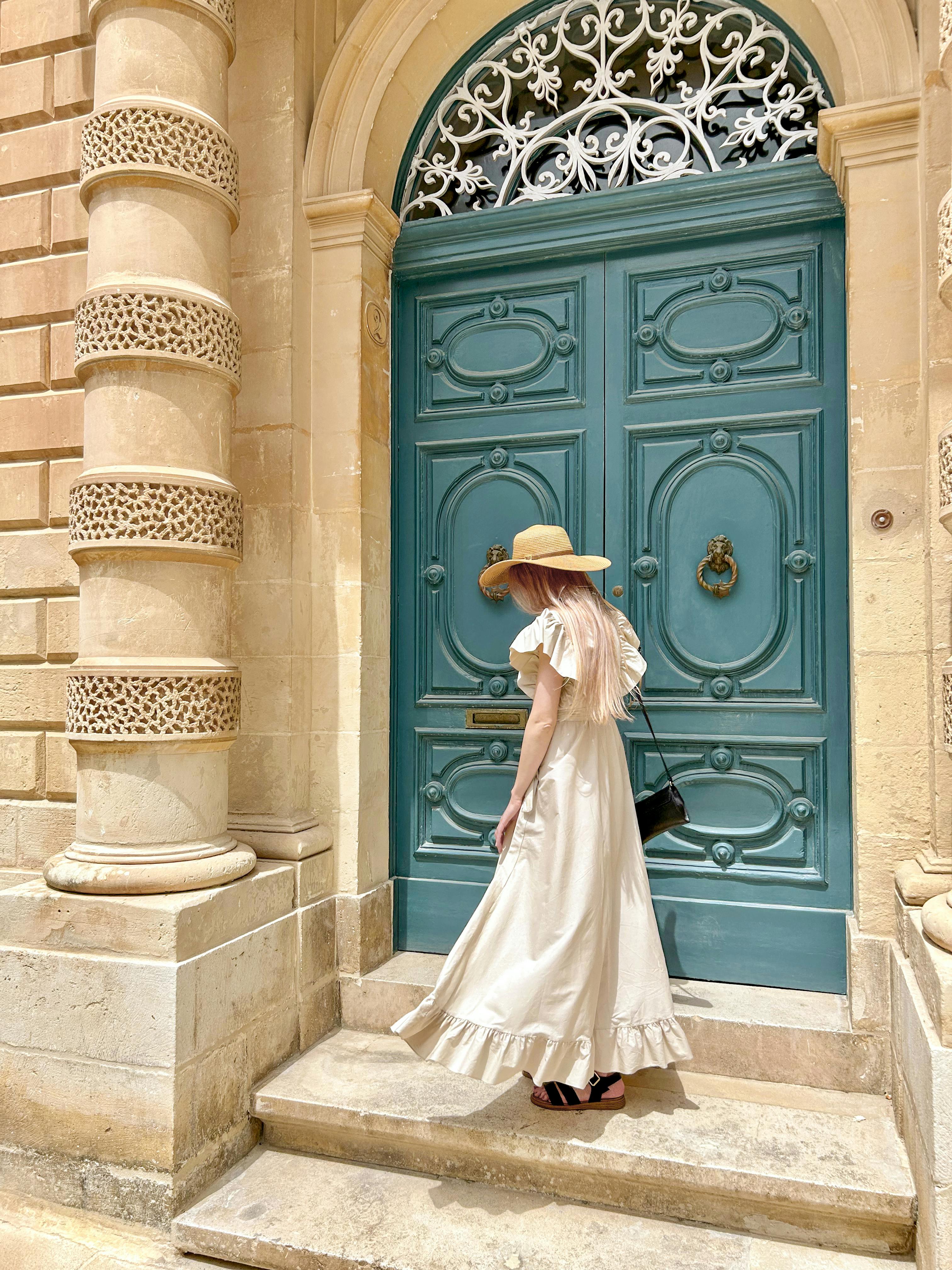 A woman in a white dress and hat stands gracefully at a vintage blue door in Mdina, Malta.