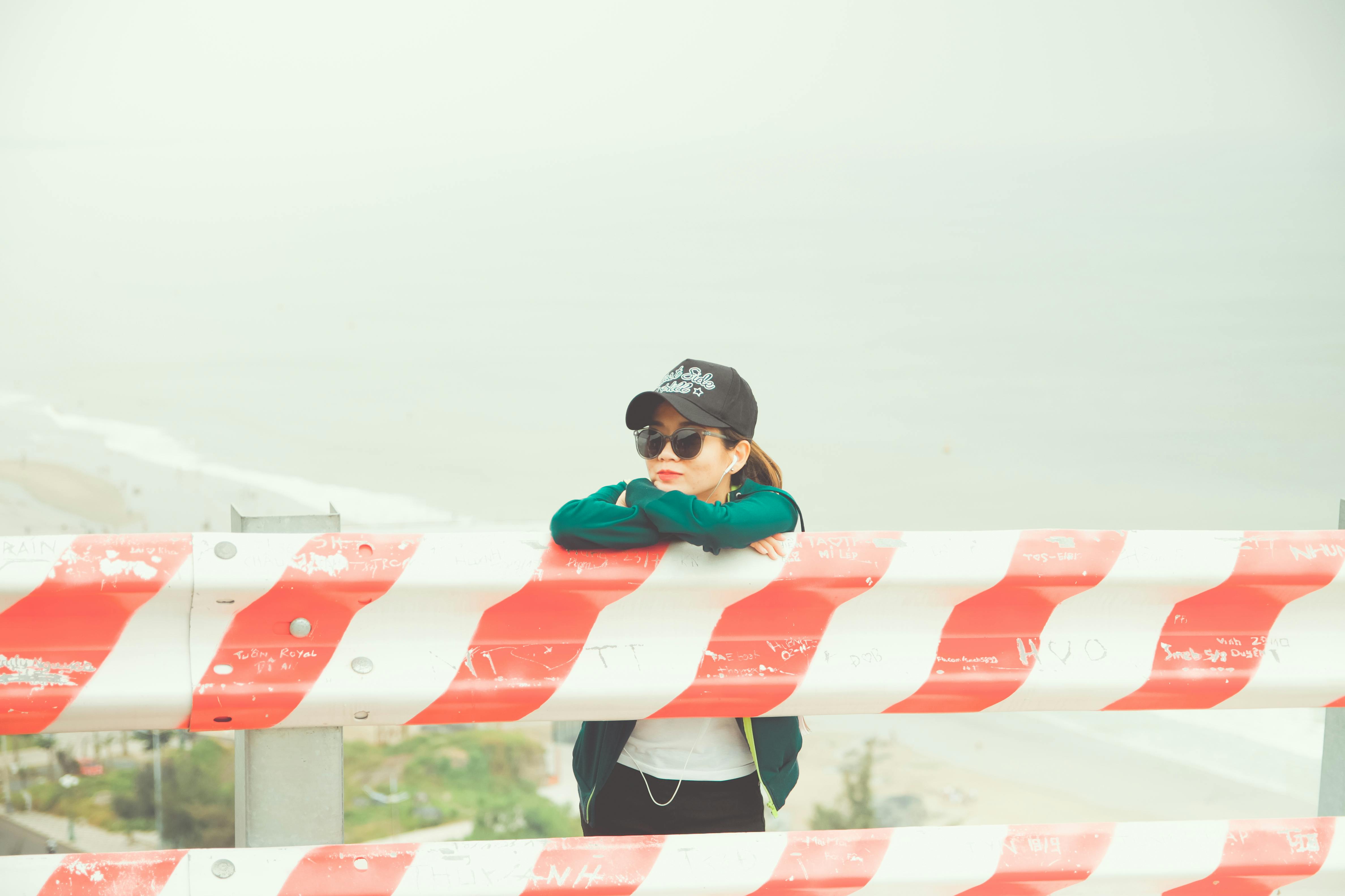 雾气弥漫的海边，一个女人戴着太阳镜和帽子，靠在红白相间的屏障上