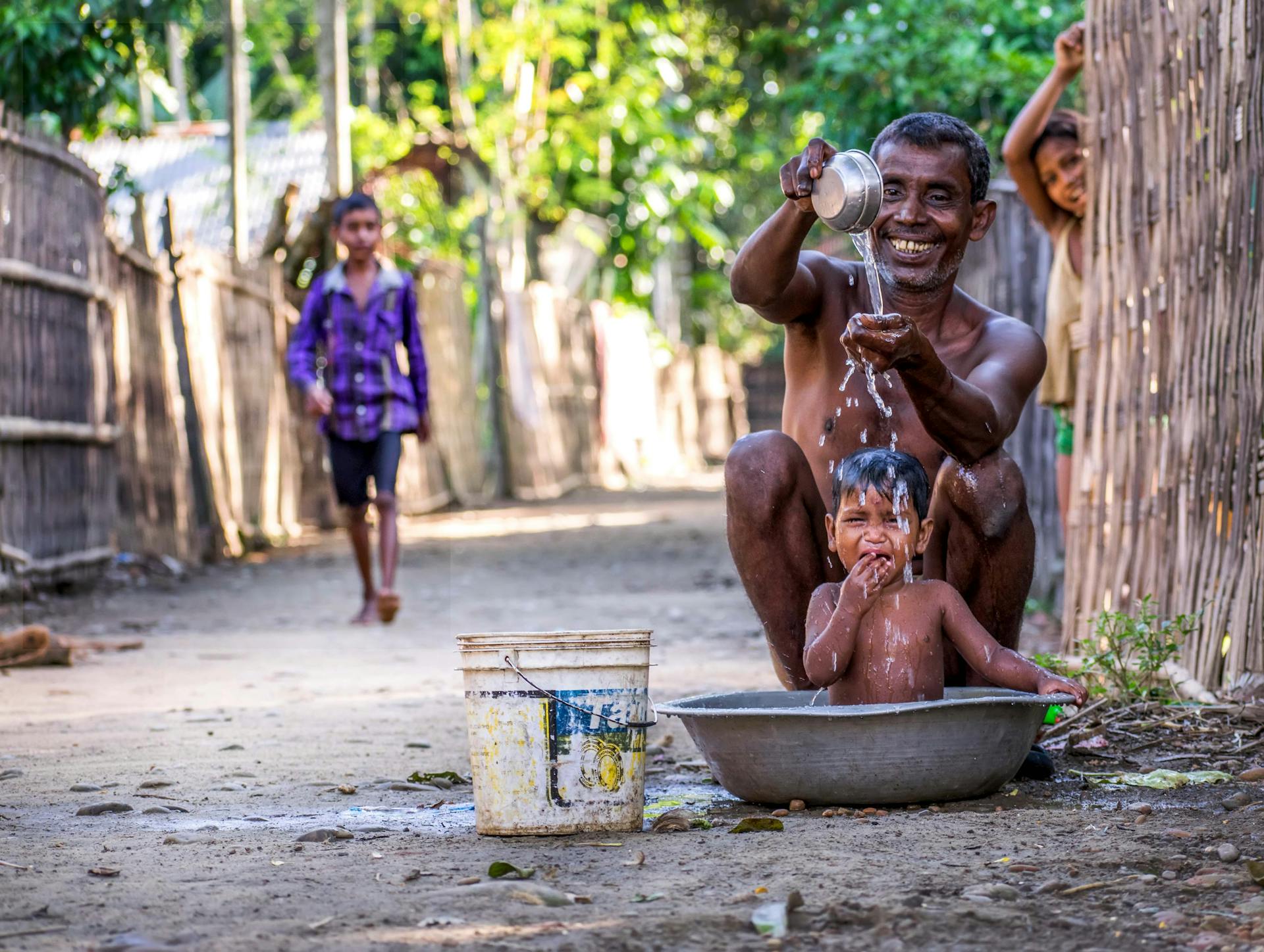 A father joyfully bathes his child in a rural Indian village setting, representing warmth and family bonding.