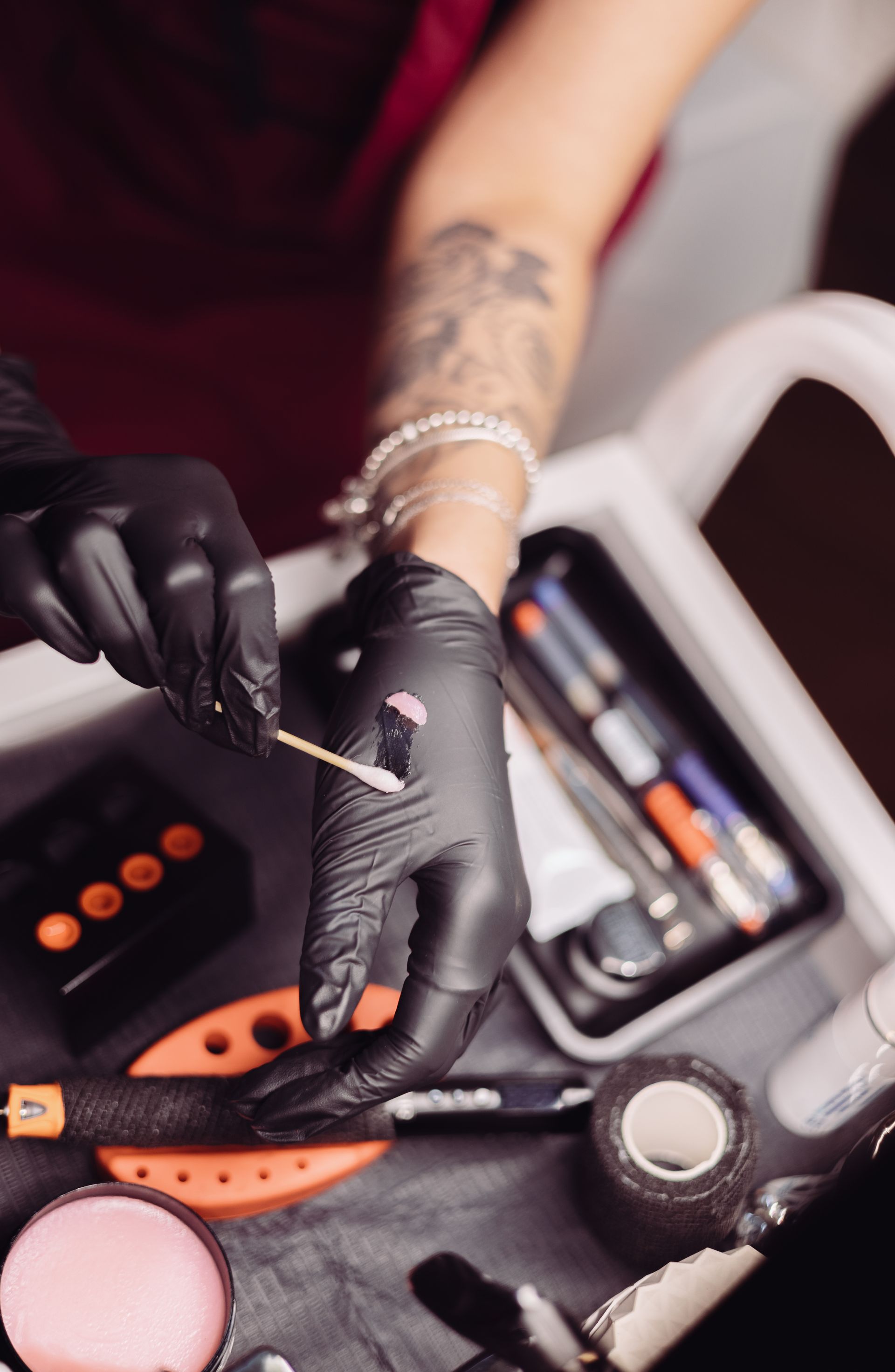 Black-gloved hands use a cotton swab near tattoo equipment and supplies, with a tattooed forearm visible.