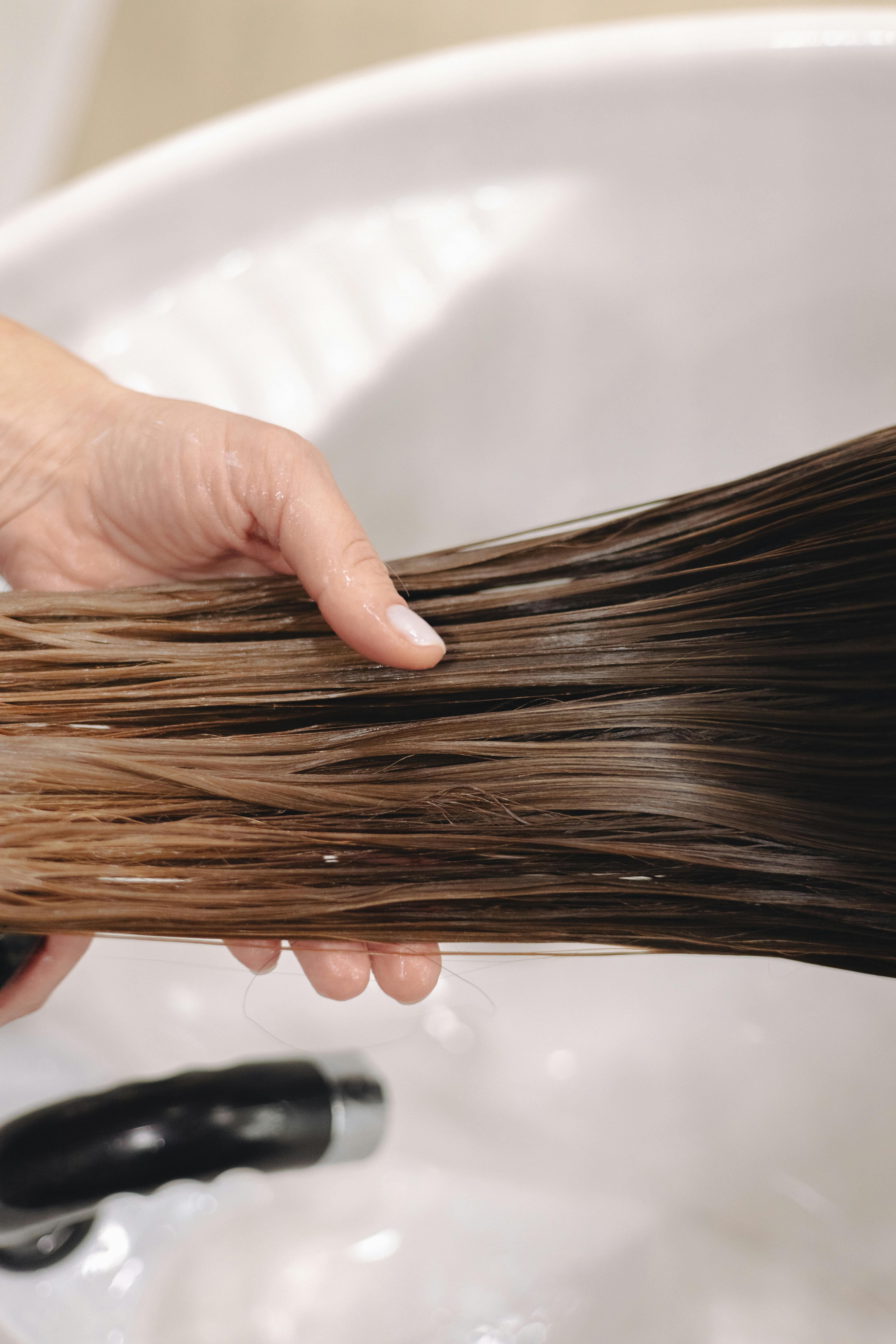 Close-up of hand holding long, wet brown hair over a white ceramic sink, emphasizing hair care.