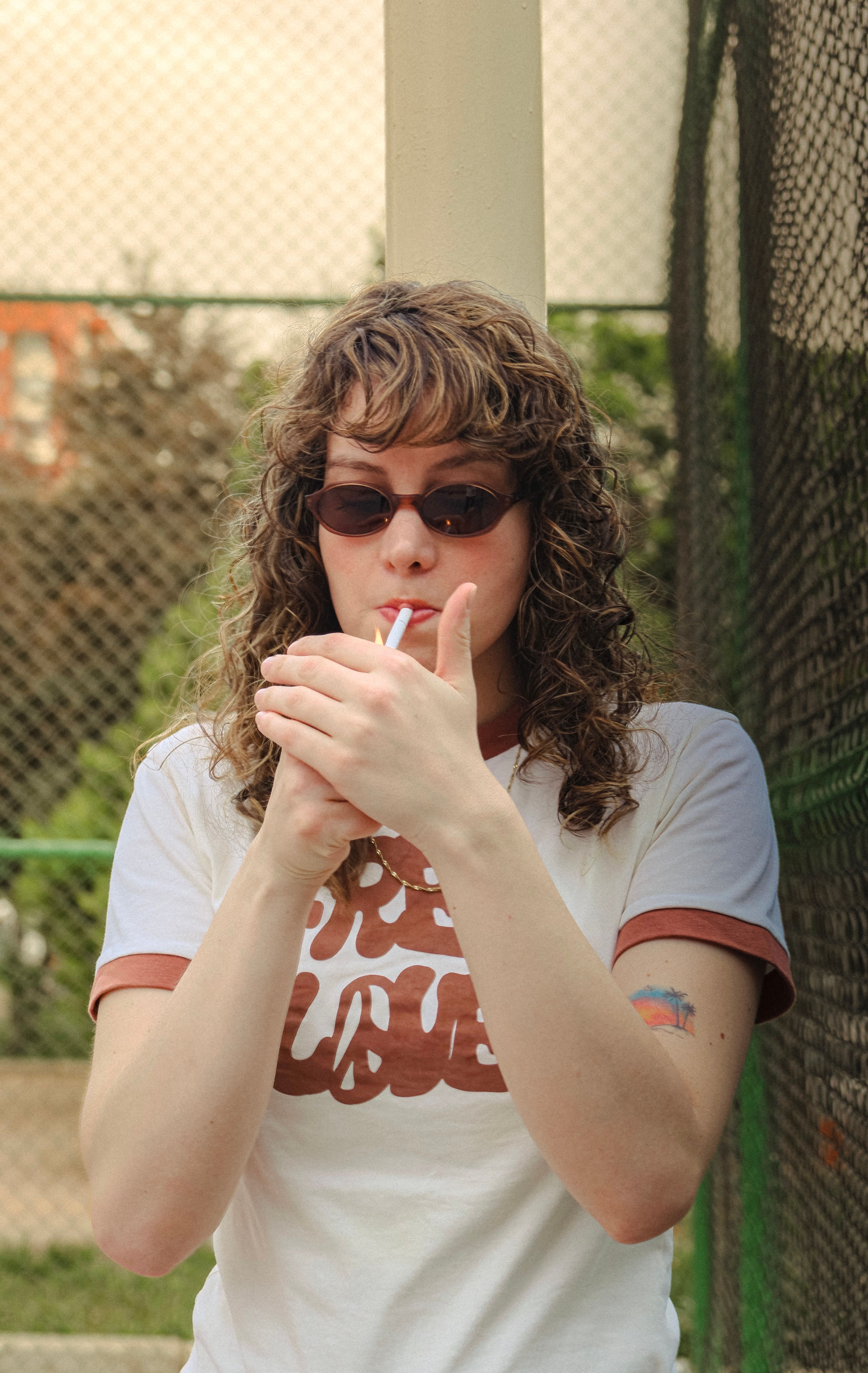 Young woman lighting a cigarette outdoors in Ankara, wearing sunglasses and a casual t-shirt.