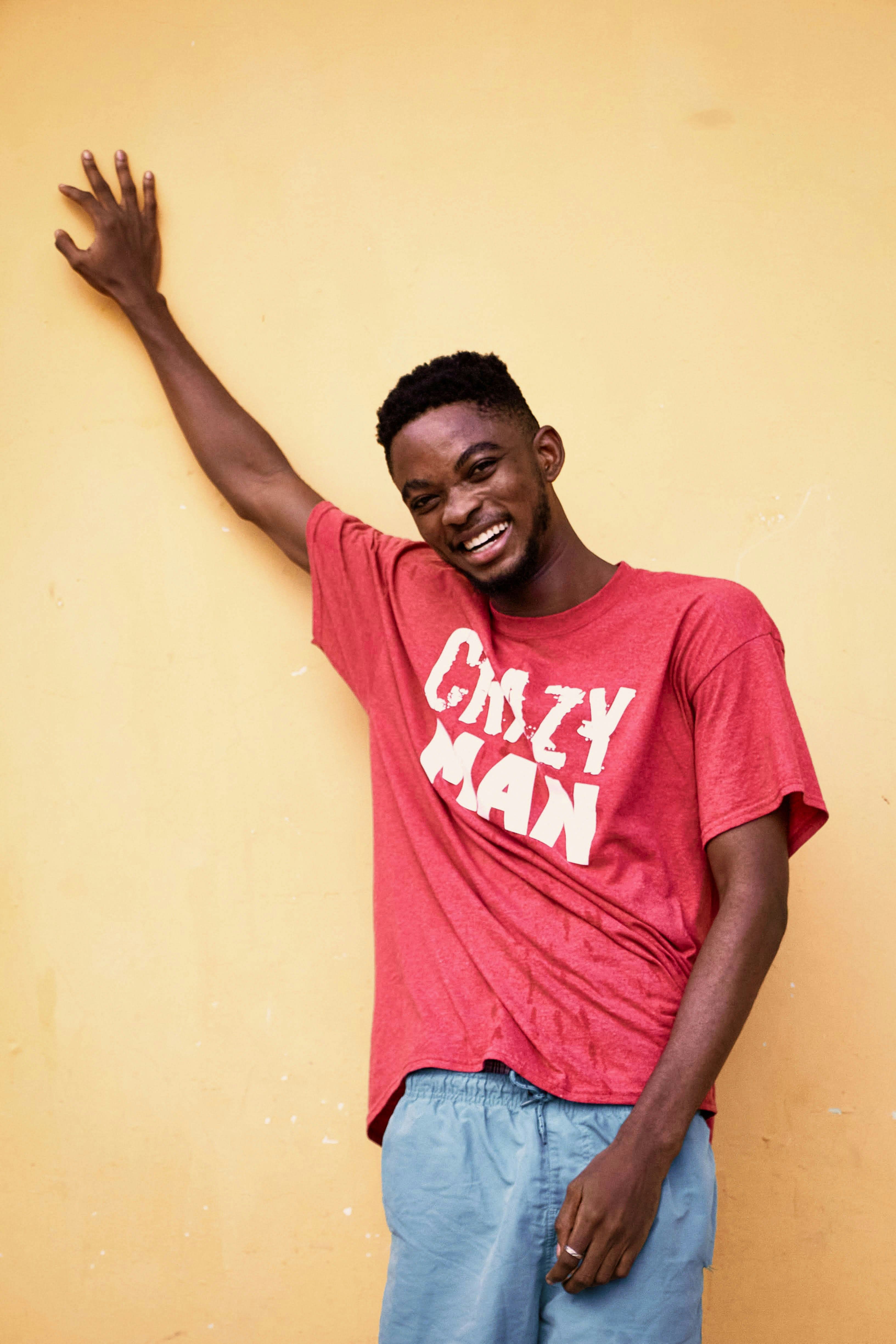A smiling man stands against a yellow wall, wearing a red t-shirt and blue jeans.