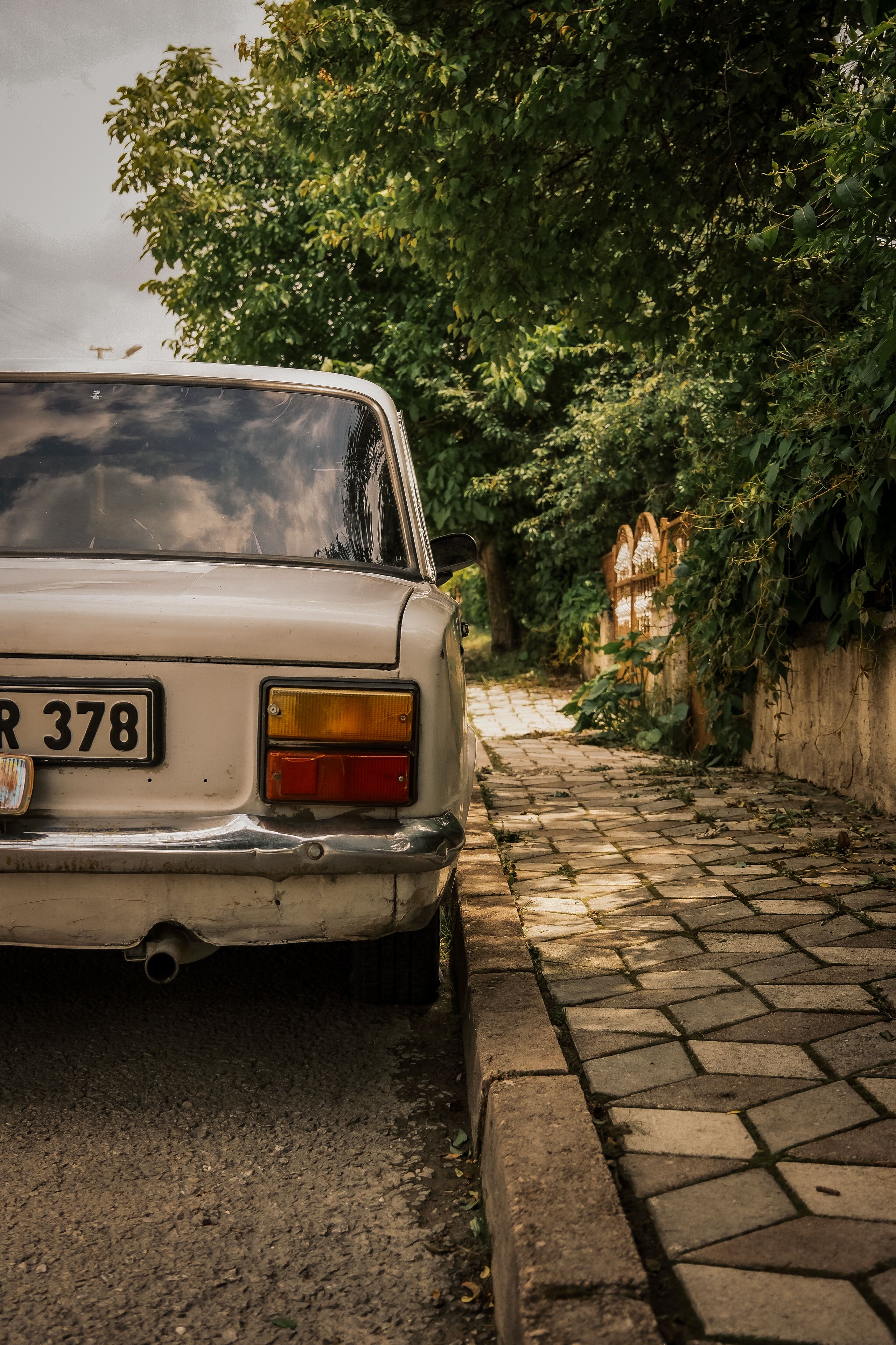 A classic car with a license plate, parked on an empty city street next to a sidewalk and green foliage.
