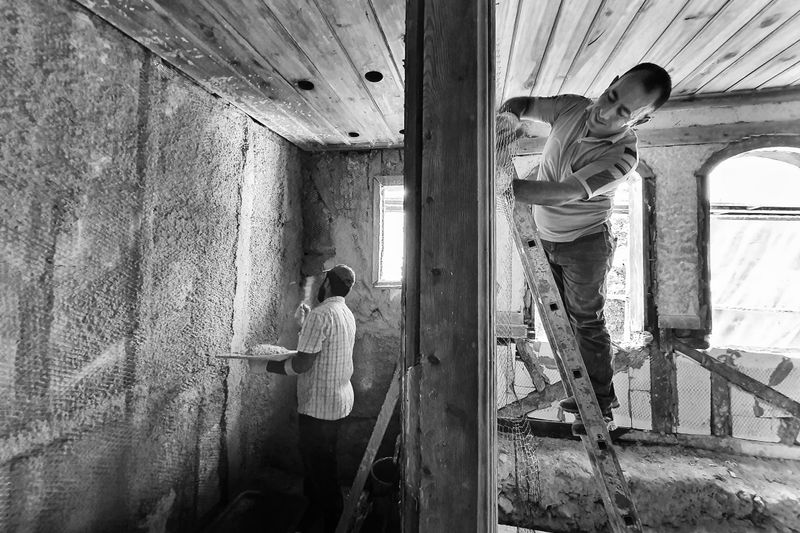 Two men renovating a room, one on a ladder applying plaster, showcasing teamwork and effort.