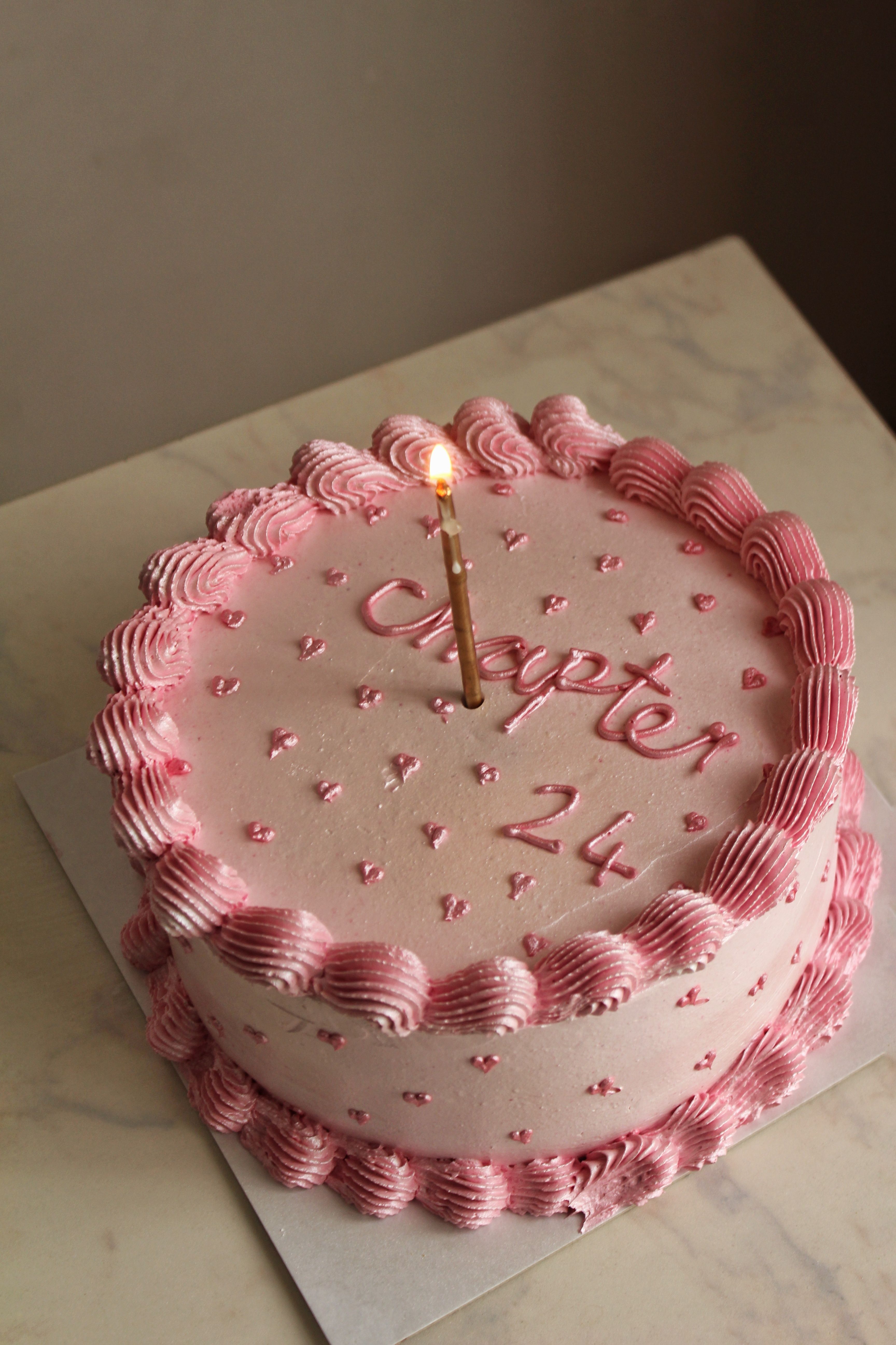 A beautifully decorated pink birthday cake with a lit candle and 'Chapter 24' written in icing.