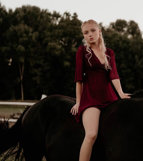A blonde woman in a burgundy dress riding a black horse
