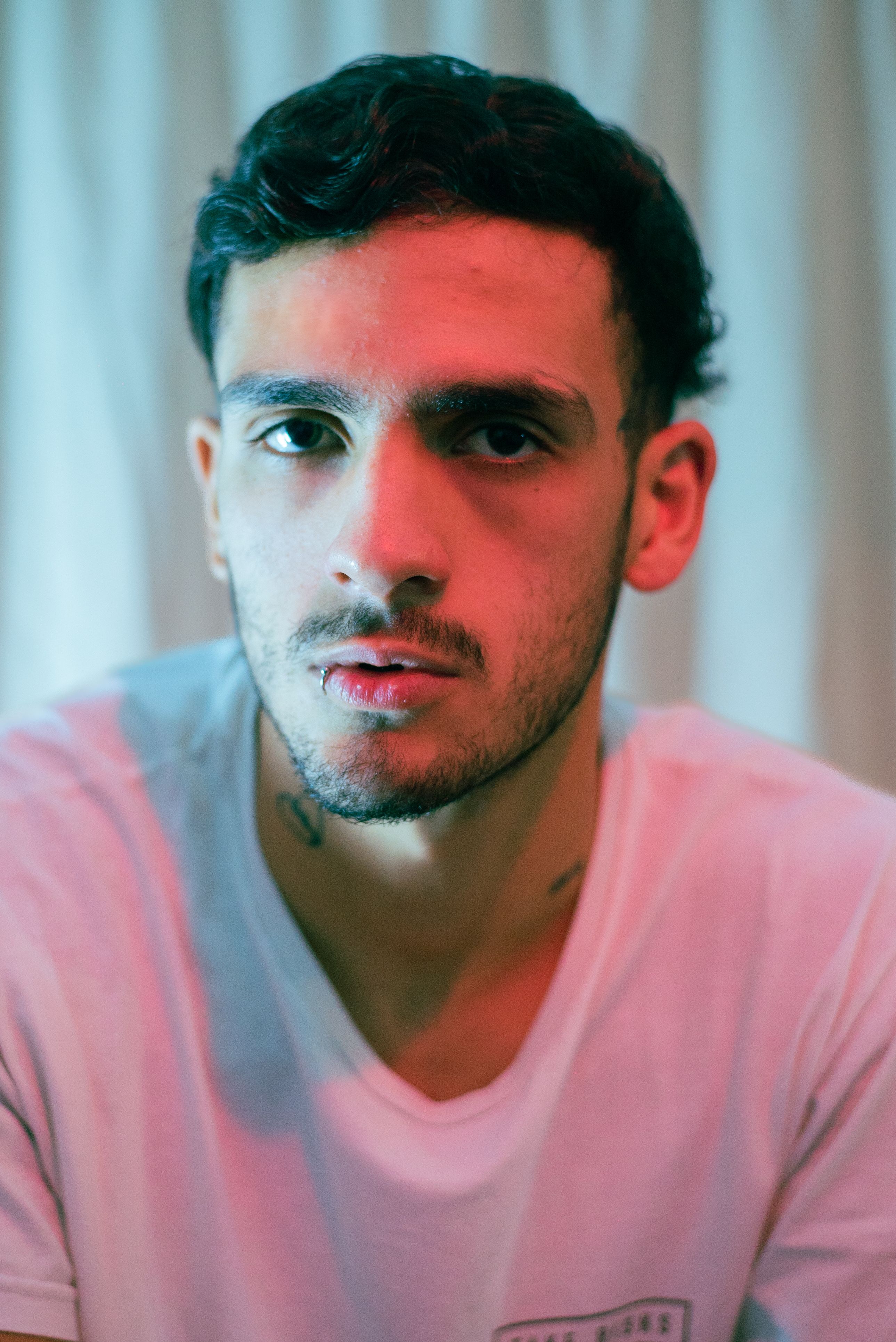 Close-up portrait of a young man with black hair and stubble wearing a white t-shirt indoors.