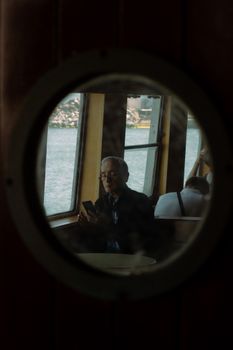 Senior man uses smartphone on a ferry with sea view through round window.