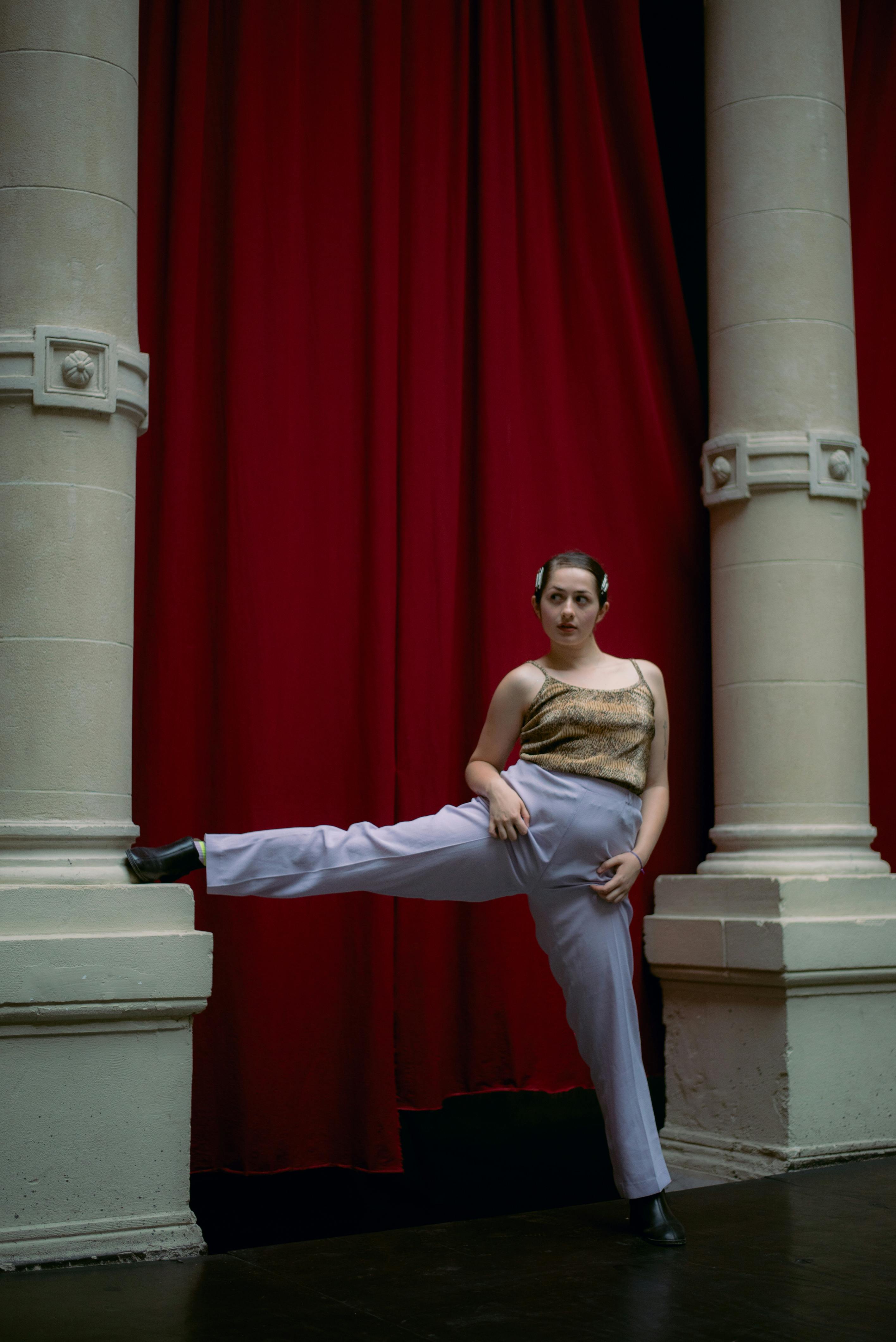 A dancer poses between theater columns with a red curtain backdrop, exuding elegance and grace.