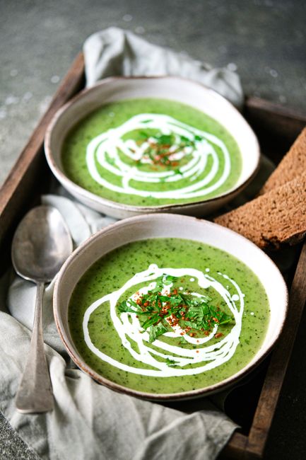 Two bowls of green soup with cream swirl and herbs, served with bread in a rustic setting.