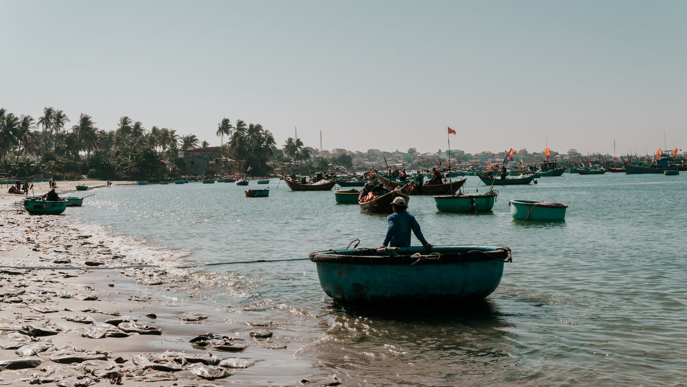Tranquil scene of a fisherman on a round boat in Mui Ne Beach, Vietnam, with palm trees and boats under a clear sky.